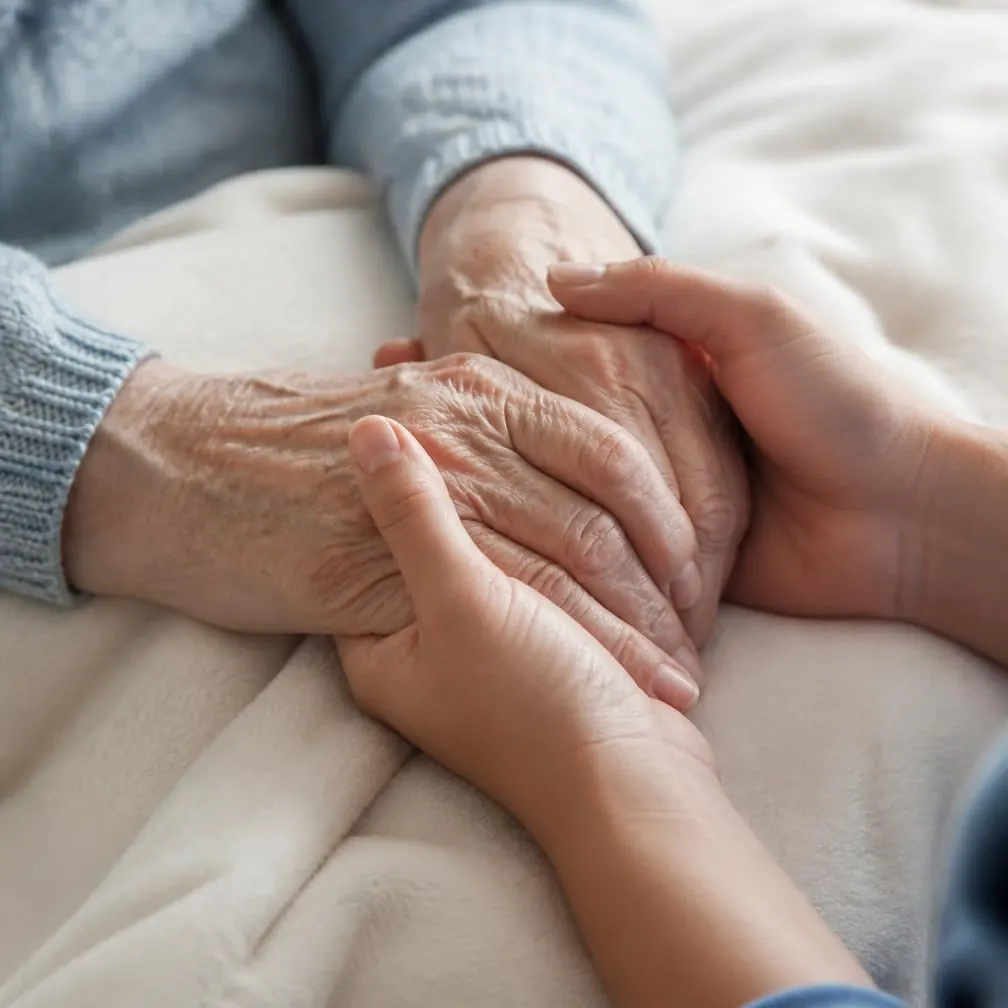 Mãos de uma cuidadora segurando as mãos de uma senhora idosa, em ambiente doméstico acolhedor